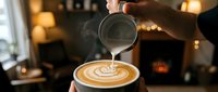 Barista pouring steamed milk creating latte art in coffee cup at cozy cafe with warm lighting and blurred background for hospitality and beverage concepts.