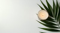 Minimalist composition with burning aromatic candle in white glass holder and fresh tropical palm leaf on light background for spa and wellness design.