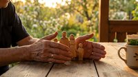 Mature man's hands cradling wooden peg dolls on rustic table with garden backdrop, symbolizing family care, protection, and togetherness for parenting and community concepts.