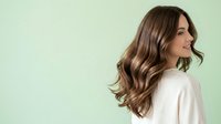 Woman with long wavy brown hair styled in loose waves, wearing a white top, photographed in profile against a soft mint green background for hair care and beauty campaigns.