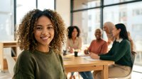 Young mixed-race businesswoman with curly hair smiling at camera in modern office meeting room with diverse team of colleagues working together.