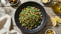 Overhead view of tabbouleh salad with parsley, tomatoes, and bulgur in black bowl on wooden table with ingredients.