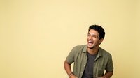 Young man with dark hair and olive skin tone, wearing a khaki button-up shirt over a dark t-shirt, smiling and looking to the side against a yellow background for portrait and lifestyle use.