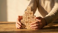 Hands protecting wooden blocks with heart symbols arranged in pyramid shape on table, representing care, love, protection and emotional support concepts.