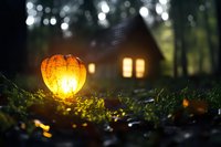 Glowing orange pumpkin lantern on mossy ground at dusk with cozy cabin windows illuminated in background creating magical autumn atmosphere.