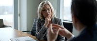 Mature woman with blonde hair and glasses listens thoughtfully during professional business meeting or job interview in modern office setting with windows.