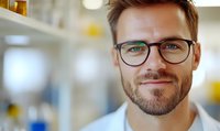 Confident male professional with beard and glasses smiling at camera in office environment. Warm natural lighting emphasizes friendly approachable expression.