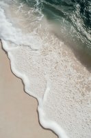 Aerial view of gentle ocean waves washing up on sandy beach with turquoise water creating white foam patterns along shoreline. Perfect for travel and nature designs.