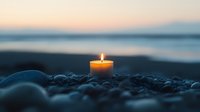 Glowing candle flame on pebble beach at dusk, soft focus candlelight against blurred ocean sunset background creates peaceful meditation atmosphere.