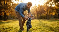 Father helping toddler son take first steps in sunny park during golden hour sunset. Family bonding moment outdoors with warm natural lighting.
