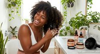 Young African American woman with curly hair applying skincare products in bathroom surrounded by green plants for natural beauty routine.