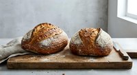 Two freshly baked artisan sourdough bread loaves with golden crusts and flour dusting on rustic wooden cutting board in bright kitchen setting.