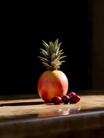 Creative fruit composition with pineapple crown attached to red apple and fresh cranberries on wooden surface in dramatic lighting against dark background.