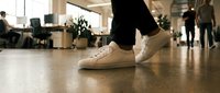 Professional walking in modern office wearing casual beige sneakers on polished floor with blurred coworkers and workspace in background.