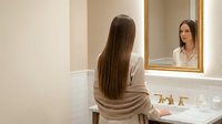 Young woman with long brown hair looking at herself in bathroom mirror while doing morning skincare routine in elegant marble vanity area.