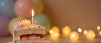 Birthday cake slice with lit candle and colorful balloons in background. Festive celebration dessert with warm bokeh lights for party occasions.