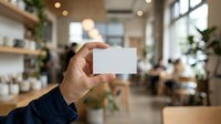 Young professional male displaying blank white business card in modern office environment with natural lighting and green plants, suitable for corporate branding and marketing materials.