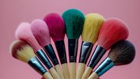 Colorful professional makeup brushes arranged in fan shape against pink background, featuring soft bristles in pink, red, yellow and black tones with wooden handles.