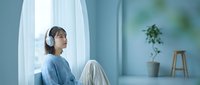 Young woman with dark hair and East Asian ethnicity wearing wireless headphones and casual blue clothing, sitting by a bright window with white curtains in a minimalist room. Suitable for lifestyle, wellness, music streaming, and remote work concepts.