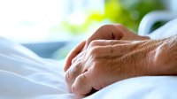 Elderly patient's hand resting on white hospital bed sheets, representing medical care, aging, and healthcare support in clinical environment.