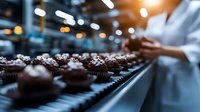 Chocolate cupcakes on conveyor belt in bakery production line with blurred pastry chef in background checking quality control. Food manufacturing process.