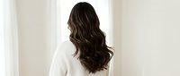Young woman with long wavy brown hair wearing white sweater standing by window with natural light for wellness and lifestyle concepts.