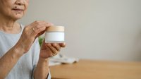 Senior woman with wrinkled hands holding white cosmetic jar with wooden lid, displaying skincare product for beauty, wellness, and anti-aging treatments.