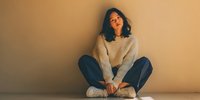 Young Asian woman in cozy sweater and jeans sitting cross-legged against beige wall, illuminated by warm natural light, conveying contemplative mood.