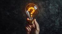 Glowing light bulb with question mark inside held by hand against dark textured background, symbolizing innovation, problem solving and creative thinking.