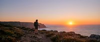 Hiker with backpack standing on rocky coastal cliff at sunrise overlooking ocean and golden sun on horizon, adventure travel and outdoor exploration concept.