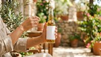 Mature man with olive skin tone holding bottle of golden wine with blank label in sunny garden setting with terracotta pots and greenery, suitable for wine marketing and lifestyle content.