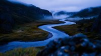 Meandering river flows through misty mountain valley at dusk, with dramatic rocky cliffs and green meadows creating moody atmospheric landscape.