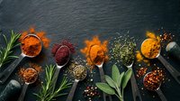 Colorful spices and herbs arranged on dark slate background with wooden spoons creating culinary cooking ingredient display for kitchen and food preparation.
