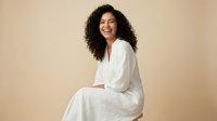 Happy young African American woman with curly hair laughing in white casual outfit against neutral beige background for lifestyle and wellness content.