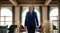 Professional mature Caucasian businesswoman with gray hair standing confidently in modern office space with exposed wooden beams and arched windows.