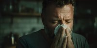 Middle-aged Caucasian man with beard blowing nose into tissue, showing signs of illness in dimly lit home environment. Concept of seasonal flu, cold, or allergies.