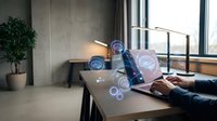 Professional working on laptop with holographic digital interface icons floating above workspace in modern office environment.