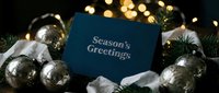 Seasons Greetings navy blue card surrounded by silver ornaments and pine branches with warm bokeh lights in festive holiday arrangement.