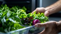 Fresh organic lettuce and red onion in meal prep container, hands arranging healthy ingredients. Dark moody lighting highlights vibrant produce for food photography.