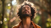 Young man with curly hair and beard basking in sunlight with eyes closed, wearing brown jacket against blurred bokeh forest background, expressing serenity and peace.