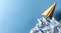 Yellow paper airplane soaring above crumpled white paper mountains against blue background, symbolizing success, achievement and rising above challenges.