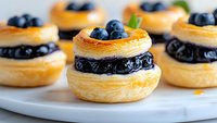 Fresh blueberry puff pastry desserts with flaky layers and sweet filling arranged on white marble plate, close up view of mini pastries garnished with berries.