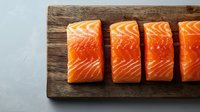Fresh raw salmon fillets arranged in row on rustic wooden cutting board, showing vibrant orange color and marbled texture. Ready for cooking preparation.
