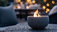 Burning flame in black stone bowl against blurred bokeh lights background creates cozy atmosphere. Decorative outdoor fire element on textured table.