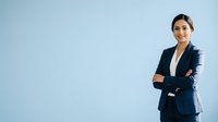 Young South Asian woman in professional navy blazer with arms crossed, smiling confidently against light blue background. Corporate portrait for business, leadership, and career concepts.