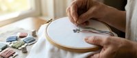 Close-up of hands embroidering delicate floral design on white fabric in wooden hoop with colorful thread spools nearby for crafting and needlework projects.
