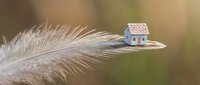 Miniature house model balanced on white feather tip against soft blurred background symbolizing fragility of home ownership and delicate real estate market.