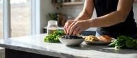Person with medium skin tone preparing fresh salad ingredients on marble countertop near window, arranging greens and vegetables for healthy meal preparation.