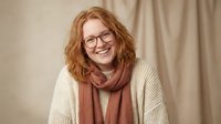Smiling middle-aged Caucasian woman with curly red hair wearing glasses and cozy sweater with scarf in warm neutral tones for lifestyle and portrait concepts.