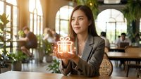 Asian woman in business attire holding glowing gift box in modern office with plants and colleagues, representing corporate rewards, employee recognition, and workplace incentives.